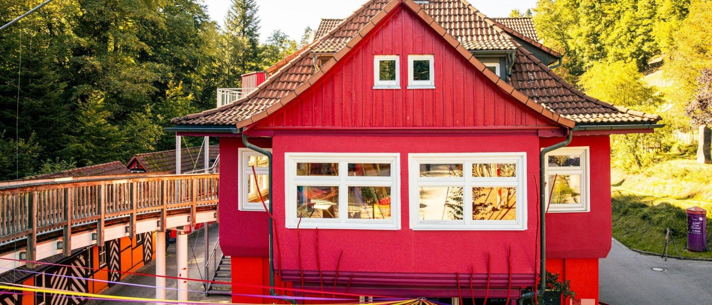 Ein rotes Gebäude mit weißen Fenstern, umgeben von grünen Bäumen. Ein Holzsteg führt am Gebäude vorbei., © Stuttgart-Marketing GmbH, Sarah Schmid