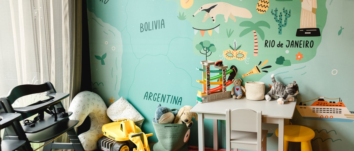 Buntes Kinderzimmer mit Spielzeug, Tisch und Stühlen vor einer Wand mit Südamerika-Karte und Illustrationen., © Rioca Stuttgart Posto 4 GmbH