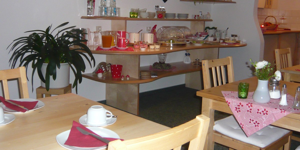 Ein gem&uuml;tlicher Fr&uuml;hst&uuml;cksraum mit Holztischen, roten Servietten und einem Buffet mit Getr&auml;nken und Speisen. Eine Pflanze steht im Raum., &copy; Hotel Hochwald
