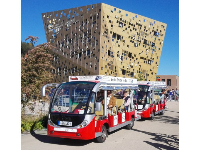 Zwei rote Elektrofahrzeuge fahren vor einem modernen, goldfarbenen Geb&auml;ude mit unregelm&auml;&szlig;igen Fenstern. Menschen sitzen in den Fahrzeugen und genie&szlig;en die Fahrt., &copy; Touristik und Marketing GmbH