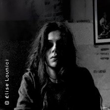 Schwarz-weiß Porträt einer Person mit langen Haaren, die in einem dunklen Raum sitzt. Im Hintergrund ist ein gerahmtes Bild an der Wand zu sehen., © links im Bild