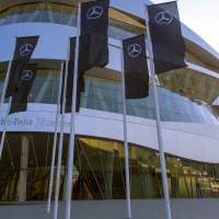 Das Mercedes-Benz Museum mit seiner modernen Architektur und mehreren Fahnen im Vordergrund. Der Himmel ist klar und blau., © SMG, Achim Mende