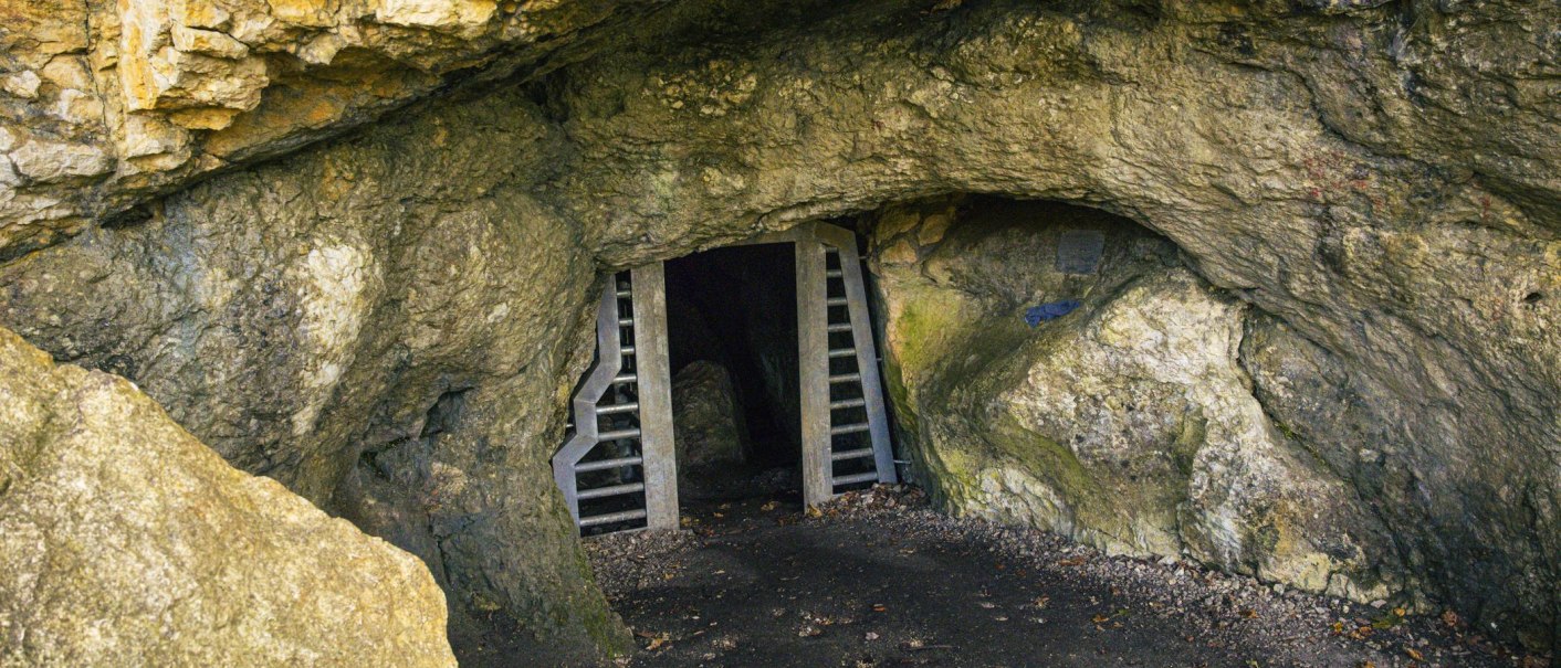 Eingang zur Schillerhöhle in Bad Urach, umgeben von Felsen. Eine Gittertür versperrt den Zugang zur dunklen Höhle., © Stuttgart-Marketing GmbH, Sarah Schmid