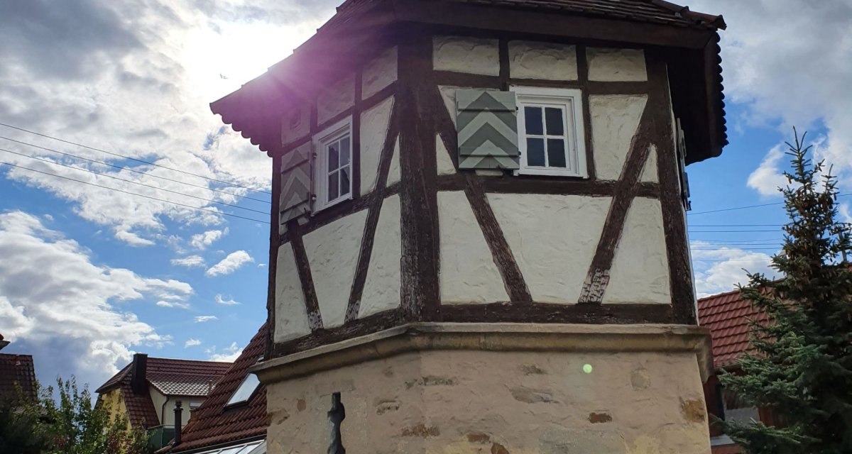 Ein Fachwerkturm mit Ziegeldach steht vor einem bew&ouml;lkten Himmel. Die Sonne scheint hinter dem Turm hervor. Im Vordergrund ist eine Hecke zu sehen.
