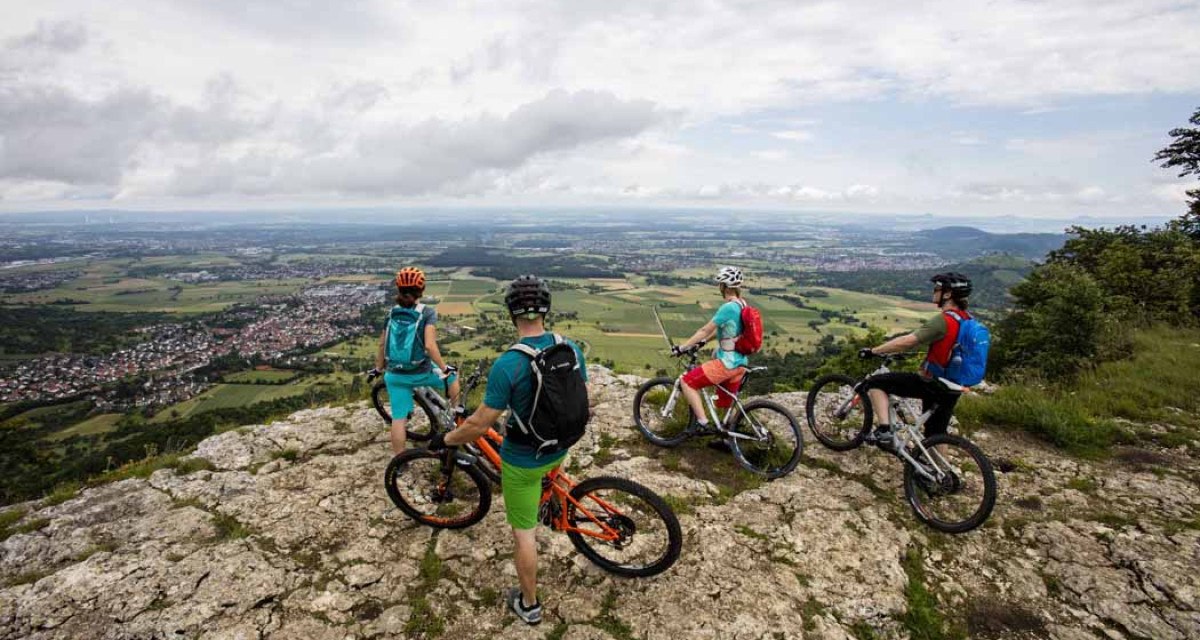 Vier Mountainbiker stehen auf einem Felsen und blicken auf eine weite Landschaft mit Feldern und Dörfern. Der Himmel ist bewölkt., © Schwäbische Alb Tourismusverband e.V.