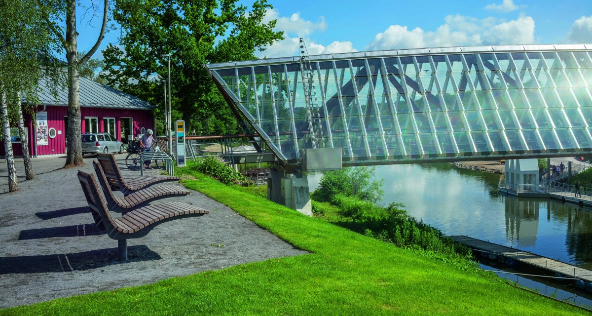 Moderne Glasbrücke in Remseck am Neckar, umgeben von grüner Landschaft und einem roten Gebäude. Sitzbänke und Fahrradständer im Vordergrund. Moderne Glasbrücke in Remseck am Neckar, umgeben von grüner Landschaft und einem roten Gebäude. Sitzbänke und Fahrradständer im Vordergrund.