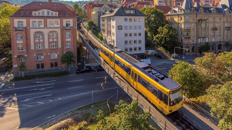 Die Zahnradbahn in Stuttgart fährt auf einer erhöhten Schiene durch eine städtische Umgebung mit historischen Gebäuden und Bäumen., © SMG, Achim Mende