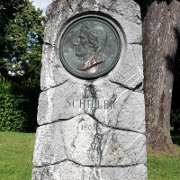 Schillerdenkmal in grüner Parkanlage mit Büschen und Bäumen - grob behauene, graue Natursteinsäule, mit bronzenem Portät-Medaillon, darunter die Inschrift "Schiller" und die Jahreszahl 1905, © Petra Natzkowski Schillerdenkmal in grüner Parkanlage mit Büschen und Bäumen - grob behauene, graue Natursteinsäule, mit bronzenem Portät-Medaillon, darunter die Inschrift "Schiller" und die Jahreszahl 1905, © Petra Natzkowski