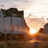 Sonnenuntergang in einer Stadtstraße mit historischen Gebäuden und Straßenbahnkabeln. Menschen überqueren die Straße, während die Sonne zwischen den Häusern untergeht., © SMG Sonnenuntergang in einer Stadtstraße mit historischen Gebäuden und Straßenbahnkabeln. Menschen überqueren die Straße, während die Sonne zwischen den Häusern untergeht., © SMG