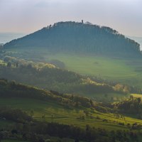 Ein bewaldeter Hügel erhebt sich in der Ferne, umgeben von grünen Feldern und Wäldern. Die Landschaft ist in sanftes Licht getaucht. Ein bewaldeter Hügel erhebt sich in der Ferne, umgeben von grünen Feldern und Wäldern. Die Landschaft ist in sanftes Licht getaucht.