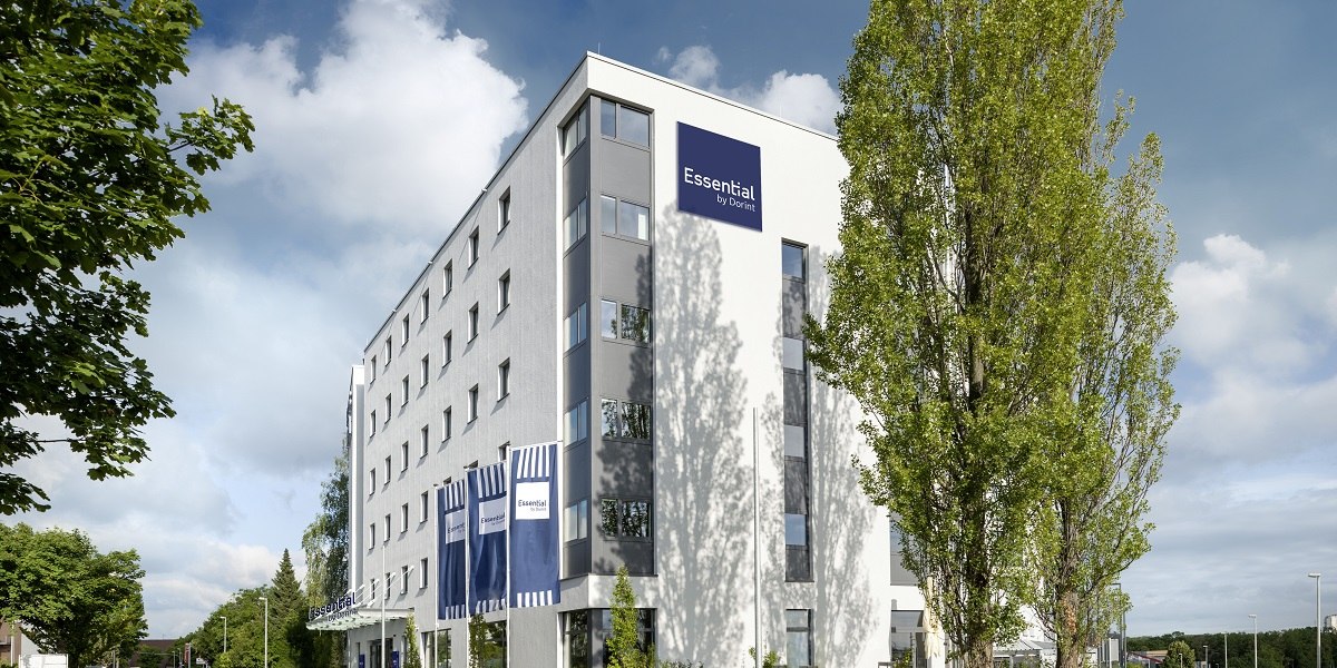 Modernes Hotelgebäude mit blauem Logo "Essential by Dorint" und Fahnen, umgeben von Bäumen und blauem Himmel., © Essential by Dorint