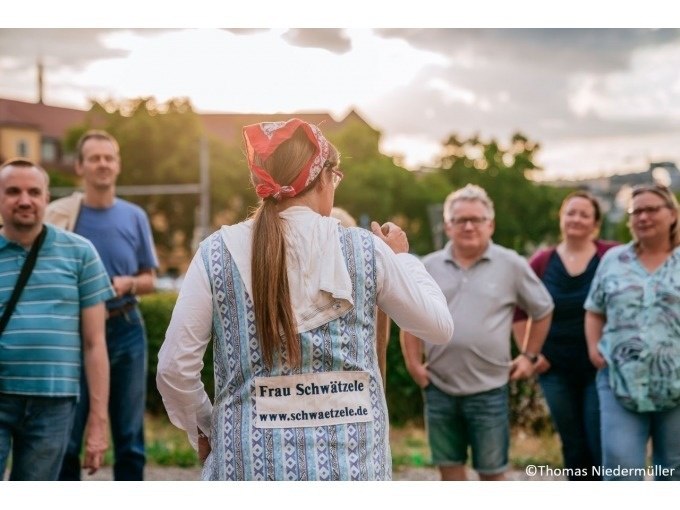 Eine Frau mit Kopftuch und Schürze spricht zu einer Gruppe von Menschen im Freien. Auf ihrer Schürze steht 'Frau Schwätzele'. Es ist sonnig., © Stuttgart Marketing GmbH Eine Frau mit Kopftuch und Schürze spricht zu einer Gruppe von Menschen im Freien. Auf ihrer Schürze steht 'Frau Schwätzele'. Es ist sonnig., © Stuttgart Marketing GmbH