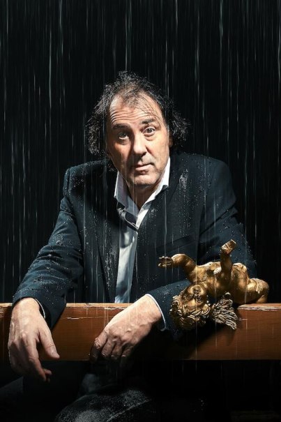 Ein Mann in einem Anzug sitzt unter Regen, mit einer goldenen Engelsfigur auf einem Tisch. Der Hintergrund ist dunkel., © Stadtverwaltung Vaihingen an der Enz Ein Mann in einem Anzug sitzt unter Regen, mit einer goldenen Engelsfigur auf einem Tisch. Der Hintergrund ist dunkel., © Stadtverwaltung Vaihingen an der Enz