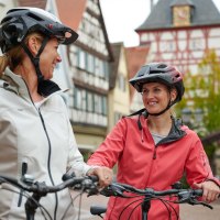 Zwei Frauen mit Helmen stehen mit Fahrrädern in einer malerischen Altstadt und lächeln sich an. Fachwerkhäuser im Hintergrund., © Kraichgau Stromberg Tourismus e.V.-Christian Ernst Zwei Frauen mit Helmen stehen mit Fahrrädern in einer malerischen Altstadt und lächeln sich an. Fachwerkhäuser im Hintergrund., © Kraichgau Stromberg Tourismus e.V.-Christian Ernst
