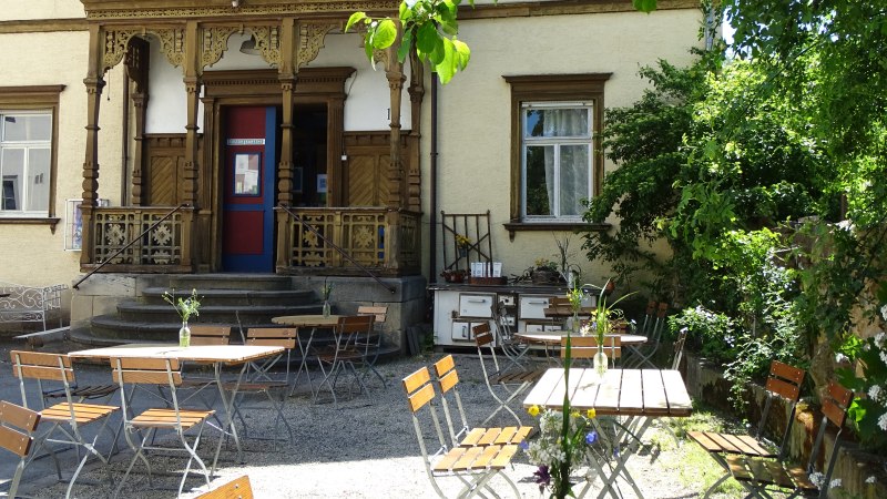 Die Alte Seegrasspinnerei mit verzierten Holzdetails und einem Au&szlig;enbereich mit Tischen und St&uuml;hlen unter B&auml;umen., &copy; Alte Seegrasspinnerei