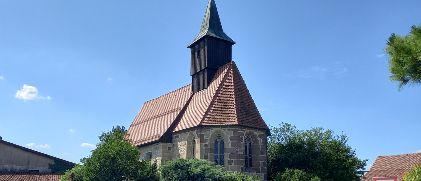 Kleine Kirche aus hellen Steinen mit dunkel verbrettertem Dachreiter auf rotbraunem Zigeldach, Blick auf den Chor mit gotischen Fenstern und die Südseite der Kirche, von Bäumen und Gebüsch umgeben, © Petra Natzkowski