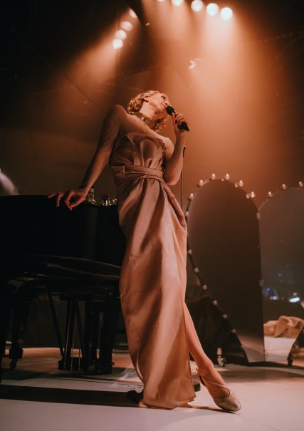 Eine Frau in einem eleganten Kleid lehnt an einem Klavier und singt in ein Mikrofon, umgeben von warmem B&uuml;hnenlicht., &copy; Ann-Marie Schwanke