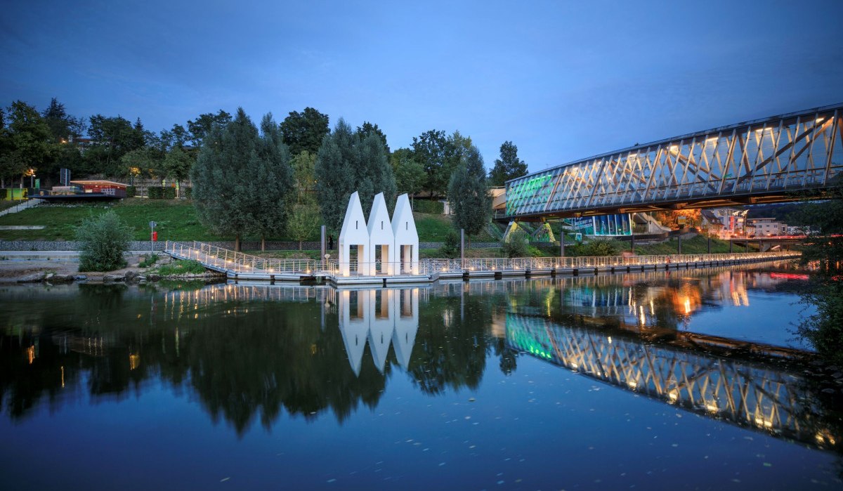 Moderne Brücke und dreieckige Gebäude am Flussufer bei Abenddämmerung, mit Spiegelung im Wasser., © Stoppel Moderne Brücke und dreieckige Gebäude am Flussufer bei Abenddämmerung, mit Spiegelung im Wasser., © Stoppel