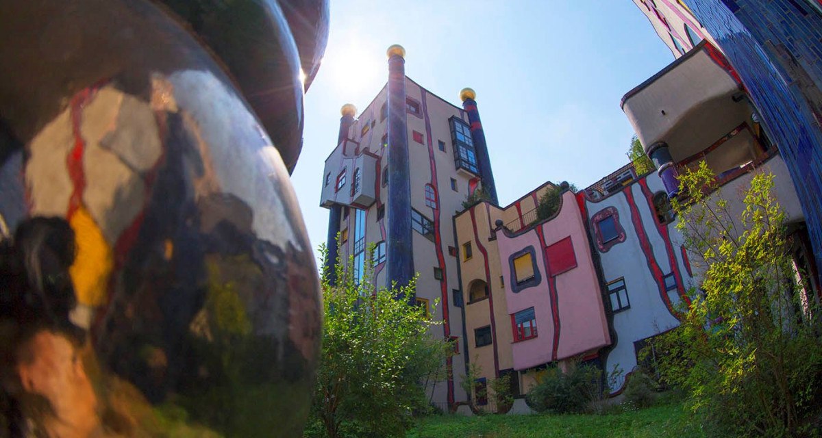 Bunte, unregelmäßige Gebäude mit runden Formen und goldenen Kuppeln im Hundertwasser-Stil, umgeben von Grünflächen., © Plochingen