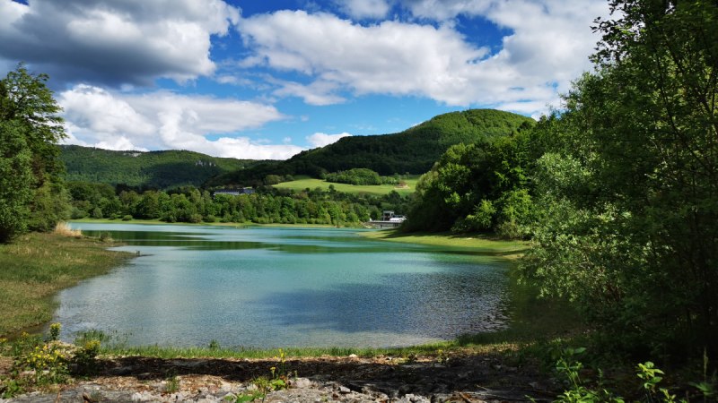 Ein idyllischer Stausee mit klarem Wasser, umgeben von grünen Hügeln und Bäumen. Der Himmel ist blau mit weißen Wolken. Ein idyllischer Stausee mit klarem Wasser, umgeben von grünen Hügeln und Bäumen. Der Himmel ist blau mit weißen Wolken.
