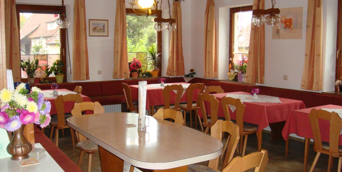 Gemütlicher Gastraum mit Holzmöbeln, roten Tischdecken und Blumen. Große Fenster mit Vorhängen lassen viel Licht herein.