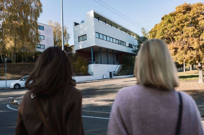 Zwei Personen stehen vor einem modernen Gebäude in der Weissenhofsiedlung, umgeben von herbstlichen Bäumen., © Stuttgart-Marketing GmbH, Ingolf Pompe