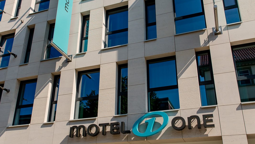 Moderne Fassade eines Gebäudes mit Motel One Logo und blauer Fahne, die im Wind weht. Klare Linien und große Fenster prägen das Design., © Motel One Stuttgart-Bad Cannstatt