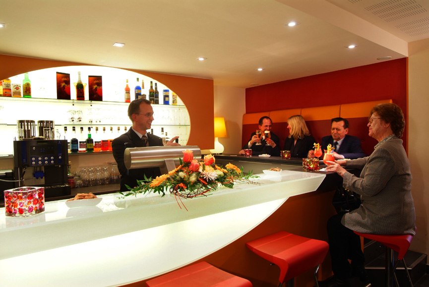 Eine moderne Bar mit Gästen, die an einem weißen Tresen sitzen. Ein Barkeeper bedient sie. Die Bar ist mit Blumen dekoriert., © Römerhof Hotelbetriebs GmbH