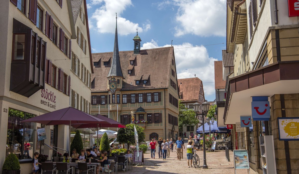 Belebte Fußgängerzone mit historischen Gebäuden, Café und Menschen unter Sonnenschirmen. Im Hintergrund ein Gebäude mit Turm und Uhr., © Stuttgart Marketing GmbH Achim Mende