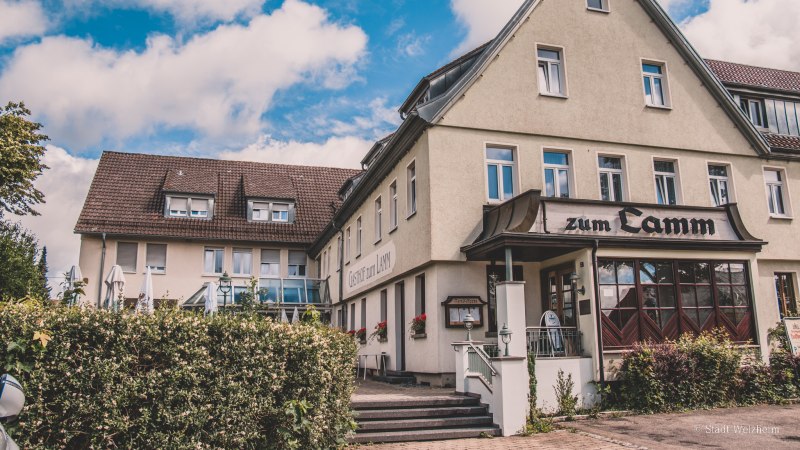 Ein traditionelles Gasthaus mit der Aufschrift 'zum Lamm', umgeben von bl&uuml;henden Str&auml;uchern und unter einem blauen Himmel mit wei&szlig;en Wolken., &copy; Stadt Welzheim