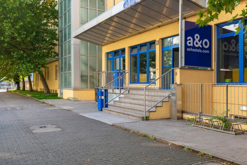 Gelbes Gebäude mit blauen Fenstern, Treppe und Vordach. Beschriftung 'a&o' und 'aohostels.com'. Baum und Gehweg im Vordergrund., © A&O Hostels Marketing GmbH