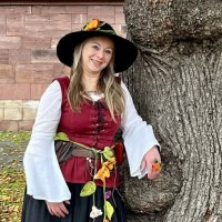 Eine Frau in mittelalterlicher Kleidung mit Blumen am Gürtel lehnt lächelnd an einem großen Baum vor einer Steinmauer., © Stadtverwaltung Weil der Stadt