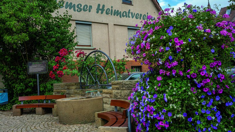 Blumenreicher Platz mit Brunnen und B&auml;nken vor einem Geb&auml;ude des Heimatvereins in Remseck am Neckar.
