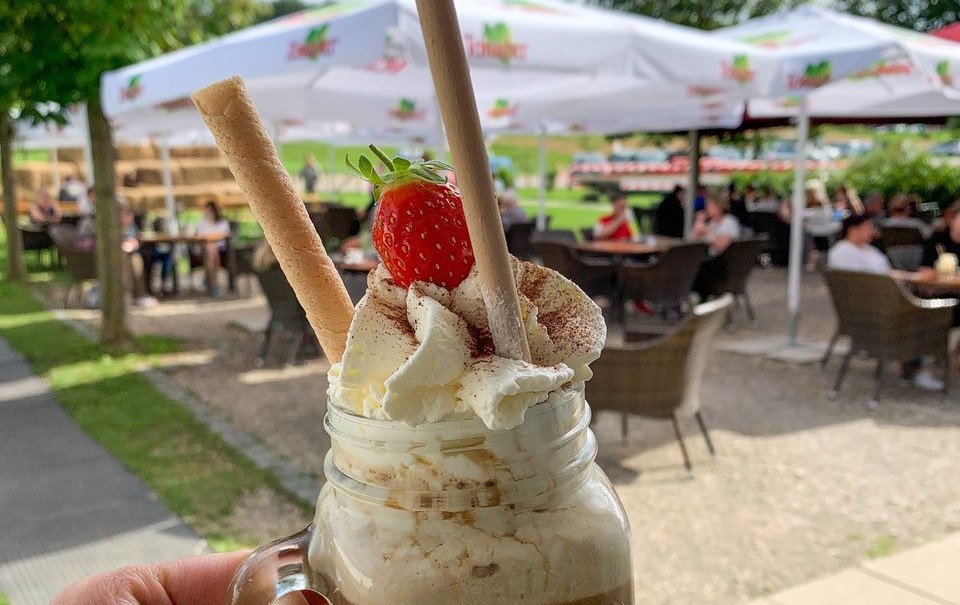 Eiskaffee mit Sahne und Erdbeere in einem Glas, gehalten vor einem Café mit Sonnenschirmen und Gästen im Freien., © Engel/ Rammerthof