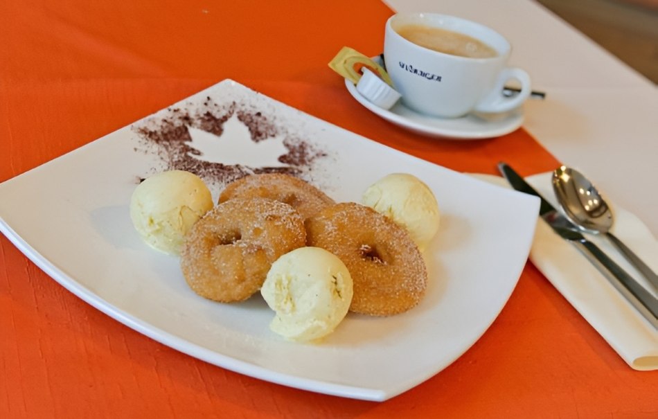 Drei Donuts mit Vanilleeiskugeln auf einem Teller, dekoriert mit Kakaopulver. Im Hintergrund eine Tasse Kaffee auf einem Tisch mit orangefarbener Tischdecke., © Hotel Neuwirtshaus