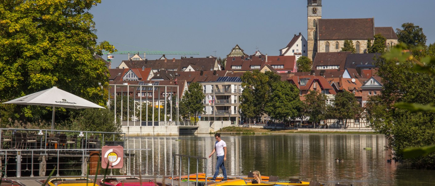 Ein See mit Tretbooten im Vordergrund, dahinter eine Kirche und Häuser in Böblingen. Ein Mann geht auf dem Steg., © Stuttgart-Marketing GmbH, Achim Mende