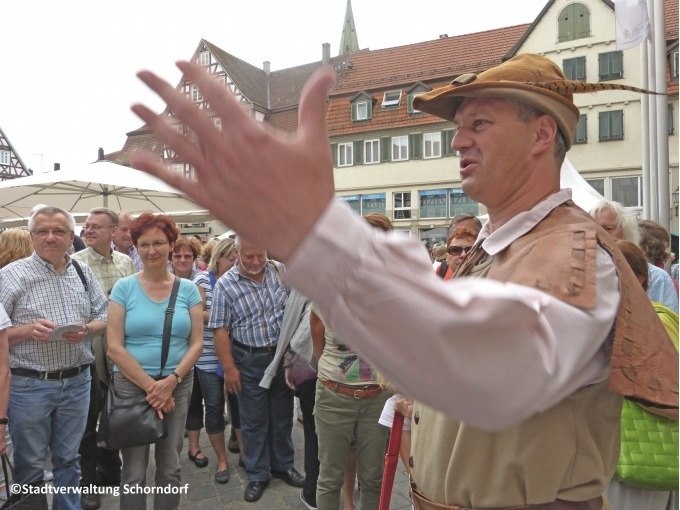 Ein Mann in historischer Kleidung spricht zu einer Gruppe von Menschen auf einem Platz in Schorndorf. Im Hintergrund sind Fachwerkhäuser zu sehen., © Eigenbetrieb Tourismus und Citymanagement Schorndorf Ein Mann in historischer Kleidung spricht zu einer Gruppe von Menschen auf einem Platz in Schorndorf. Im Hintergrund sind Fachwerkhäuser zu sehen., © Eigenbetrieb Tourismus und Citymanagement Schorndorf