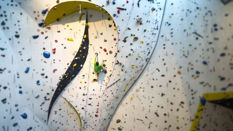 Ein Kletterer in grüner Kleidung erklimmt eine hohe Indoor-Kletterwand mit bunten Griffen im DAV Kletter- und Boulderzentrum Schwaben., © Rockerei