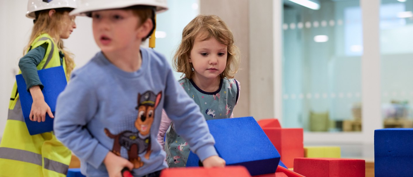 Kinder spielen auf einer Indoor-Baustelle mit bunten Bauklötzen. Sie tragen Bauhelme und Sicherheitswesten. Im Hintergrund sind moderne Räume zu sehen., © Julia Ochs Kinder spielen auf einer Indoor-Baustelle mit bunten Bauklötzen. Sie tragen Bauhelme und Sicherheitswesten. Im Hintergrund sind moderne Räume zu sehen., © Julia Ochs