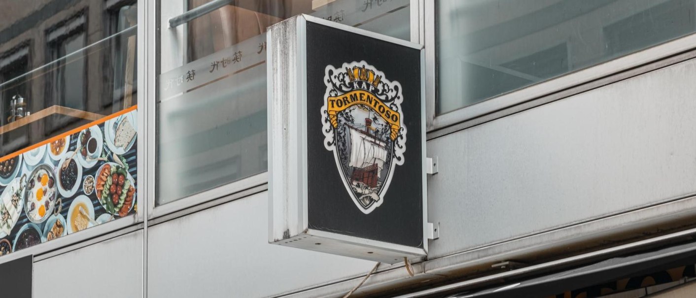 Ein Schild mit dem Logo von Kap Tormentoso hängt an einem modernen Gebäude. Es zeigt ein Segelschiff und dekorative Elemente., © Stuttgart Marketing GmbH, Sarah Schmid Ein Schild mit dem Logo von Kap Tormentoso hängt an einem modernen Gebäude. Es zeigt ein Segelschiff und dekorative Elemente., © Stuttgart Marketing GmbH, Sarah Schmid