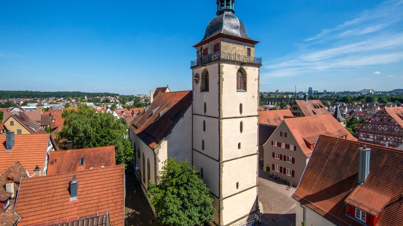 Die Evangelische Stadtkirche Bietigheim-Bissingen ragt über die roten Dächer der Altstadt, umgeben von Bäumen und unter einem klaren blauen Himmel., © SMG, Achim Mende Die Evangelische Stadtkirche Bietigheim-Bissingen ragt über die roten Dächer der Altstadt, umgeben von Bäumen und unter einem klaren blauen Himmel., © SMG, Achim Mende