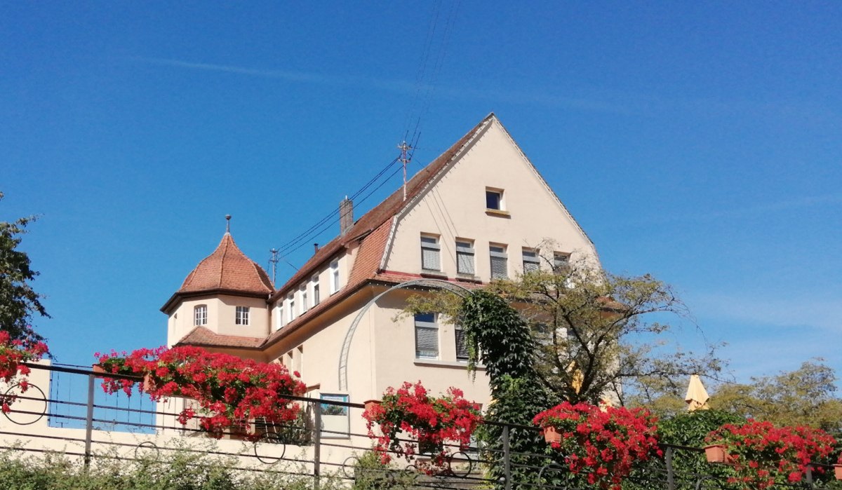 Das alte Schulhaus Bargau mit roten Dachziegeln und einem Turm. Im Vordergrund blühende rote Blumen und ein klarer blauer Himmel., © Foto: Cornelia Steinbach Das alte Schulhaus Bargau mit roten Dachziegeln und einem Turm. Im Vordergrund blühende rote Blumen und ein klarer blauer Himmel., © Foto: Cornelia Steinbach