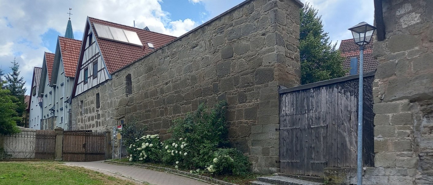 Historische Stadtmauer in Gaildorf, flankiert von Fachwerkh&auml;usern und einem gepflasterten Gehweg. Ein blauer Himmel mit Wolken bildet den Hintergrund., &copy; Petra Natzkowski