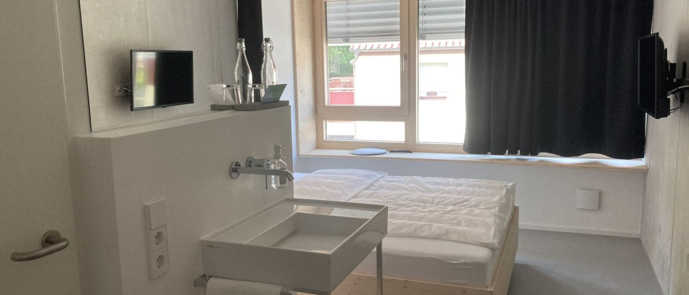 Modernes Zimmer mit Holzrahmenbett, Waschbecken, Fernseher und großem Fenster mit Vorhängen. Schlichte, minimalistische Einrichtung., © Remstal Tourismus e.V. Modernes Zimmer mit Holzrahmenbett, Waschbecken, Fernseher und großem Fenster mit Vorhängen. Schlichte, minimalistische Einrichtung., © Remstal Tourismus e.V.