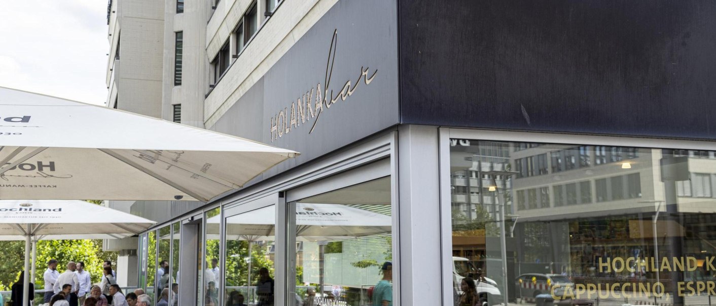 Au&szlig;enansicht der Holanka Bar mit gro&szlig;en Sonnenschirmen. Menschen sitzen und stehen im Au&szlig;enbereich. Moderne Geb&auml;ude im Hintergrund., &copy; SMG, Sarah Schmid