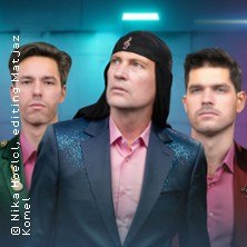 Drei M&auml;nner in eleganter Kleidung posieren vor einem blauen Hintergrund. Einer tr&auml;gt ein schwarzes Stirnband, alle schauen in verschiedene Richtungen., &copy; links im Bild