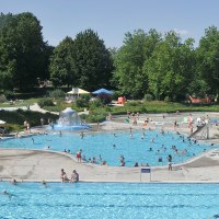Ein belebtes Freibad mit mehreren Pools, vielen Besuchern und umgeben von grünen Bäumen. Menschen schwimmen und entspannen auf Liegestühlen., © Werner Kuhnle