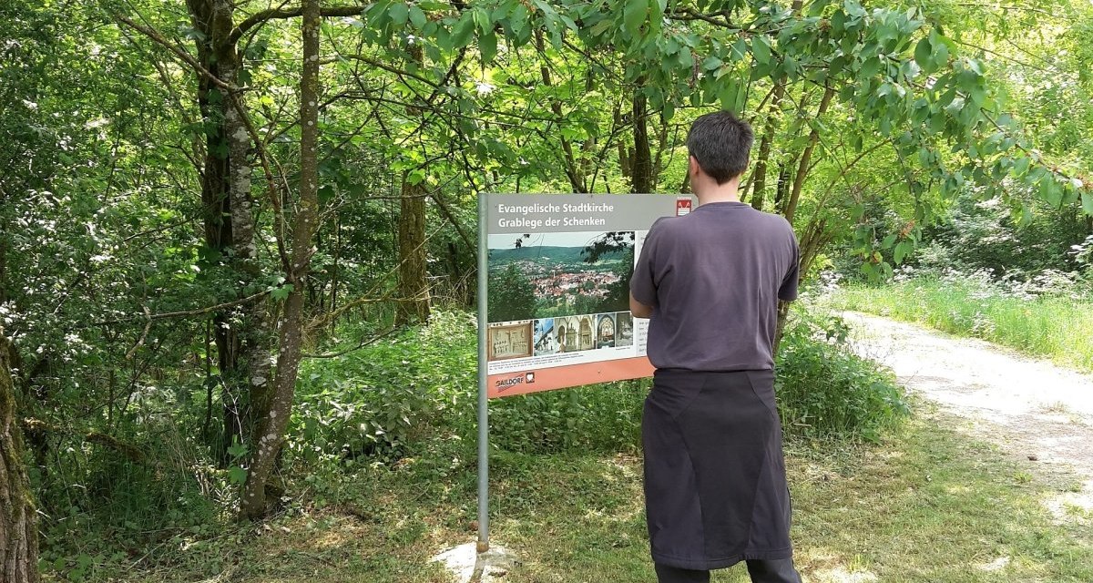Ein Mann betrachtet eine Infotafel im Wald. Die Tafel informiert über die evangelische Stadtkirche und Grablege der Schenken in Gaildorf. Umgeben von Bäumen., © Petra Natzkowski Ein Mann betrachtet eine Infotafel im Wald. Die Tafel informiert über die evangelische Stadtkirche und Grablege der Schenken in Gaildorf. Umgeben von Bäumen., © Petra Natzkowski