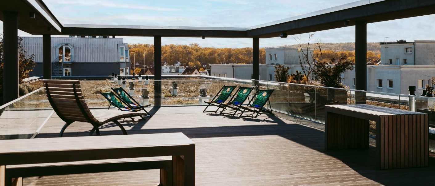 Moderne Dachterrasse mit Liegestühlen und Holzmöbeln, umgeben von Glasgeländern, mit Blick auf Gebäude und Herbstlandschaft., © Rioca Stuttgart Posto 4 GmbH