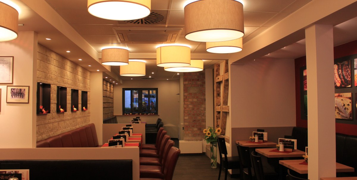 Gemütliches Restaurant mit roten Tischdecken, modernen Lampen und Wanddekorationen. Sitzplätze entlang der Wände, warme Beleuchtung.